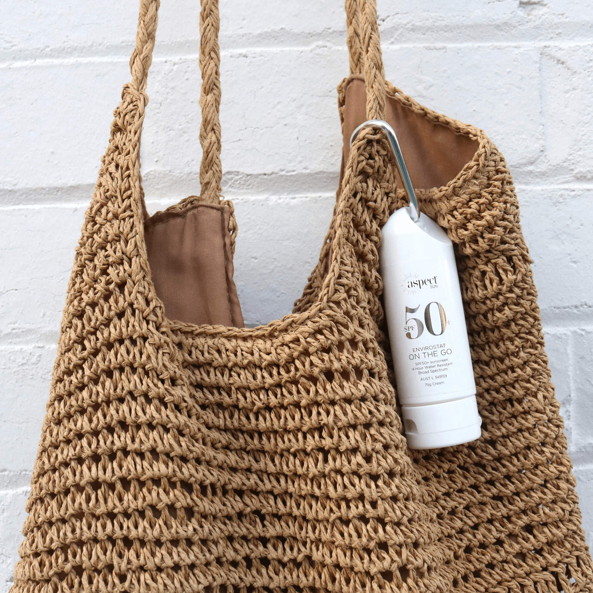 Brown crochet bag with a bottle of Aspect sun cream on a white brick wall background