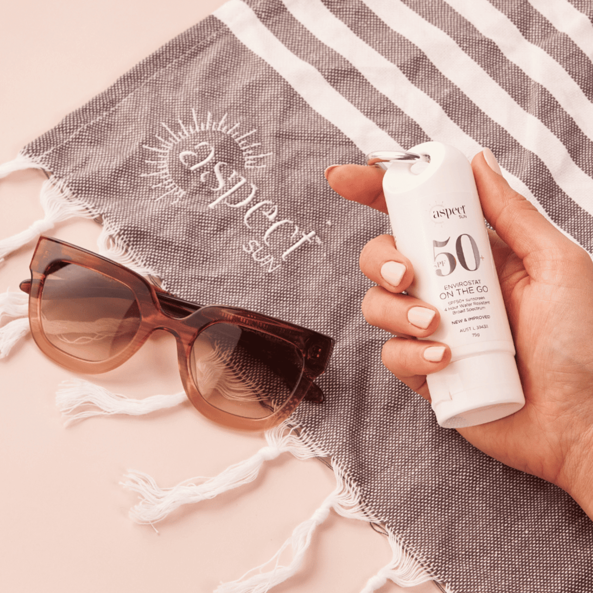 Hand holding a sunscreen bottle labeled 'aspect' with sunglasses and a towel in the background.