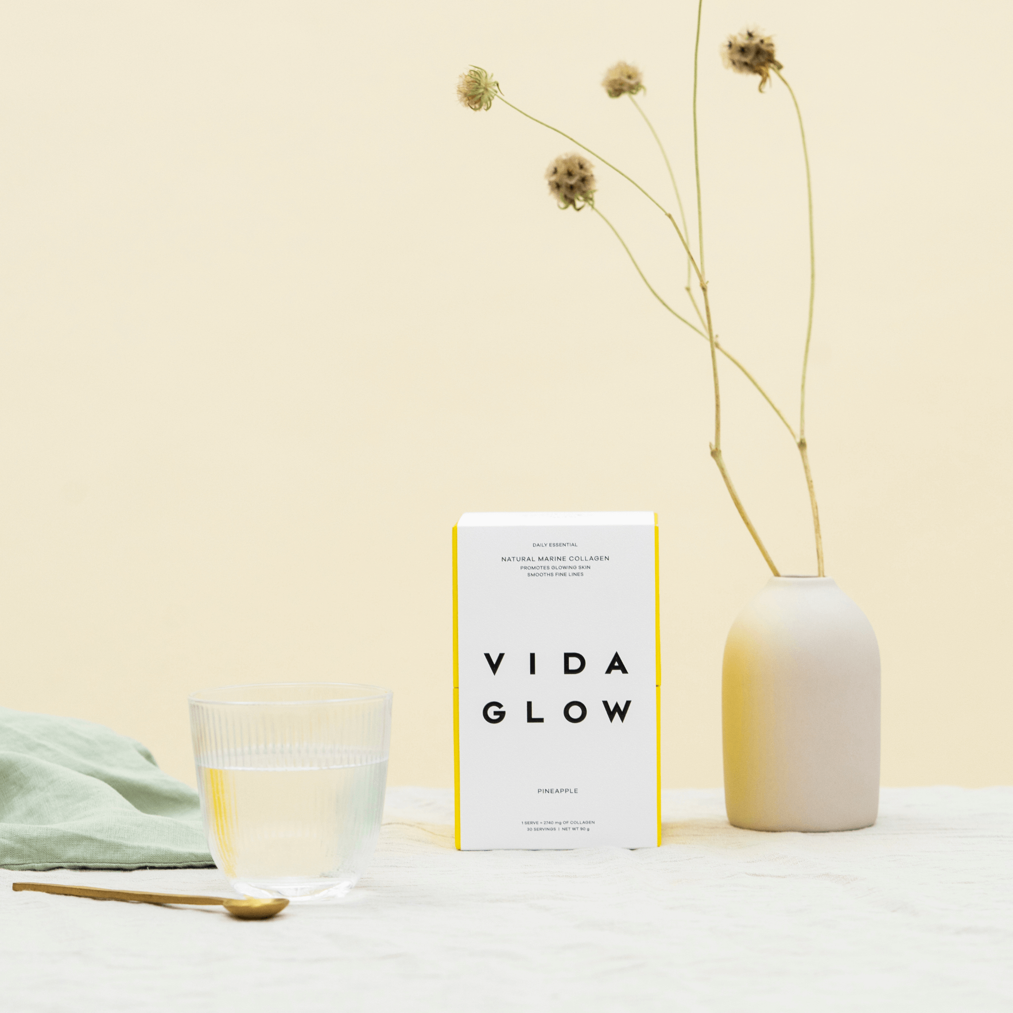 Vida Glow pineapple collagen powder on yellow background with flowers and glass of water