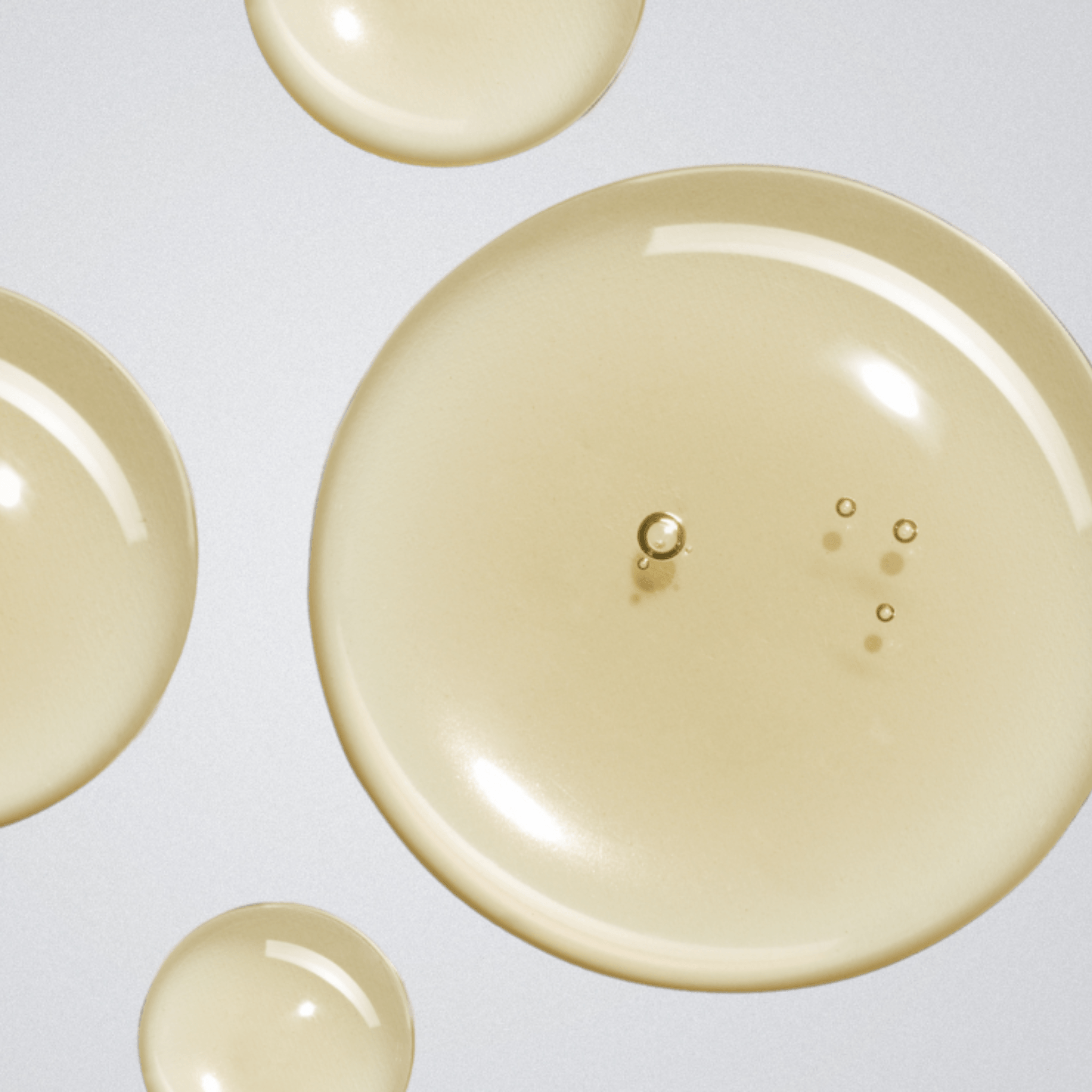 A close-up shot of several translucent, pale yellow liquid droplets of Aspect Dr Mild Clean on a light grey background