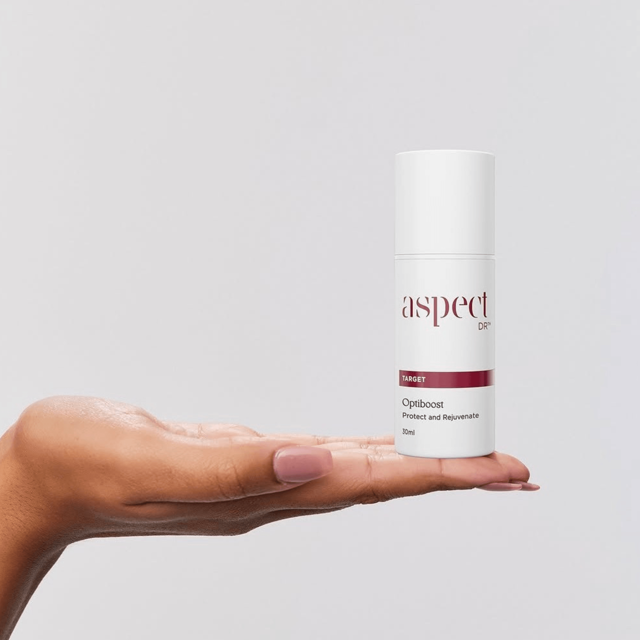 A hand holding a white cylindrical bottle of Aspect Dr Optiboost Protect and Rejuvenate serum against a light grey background.
