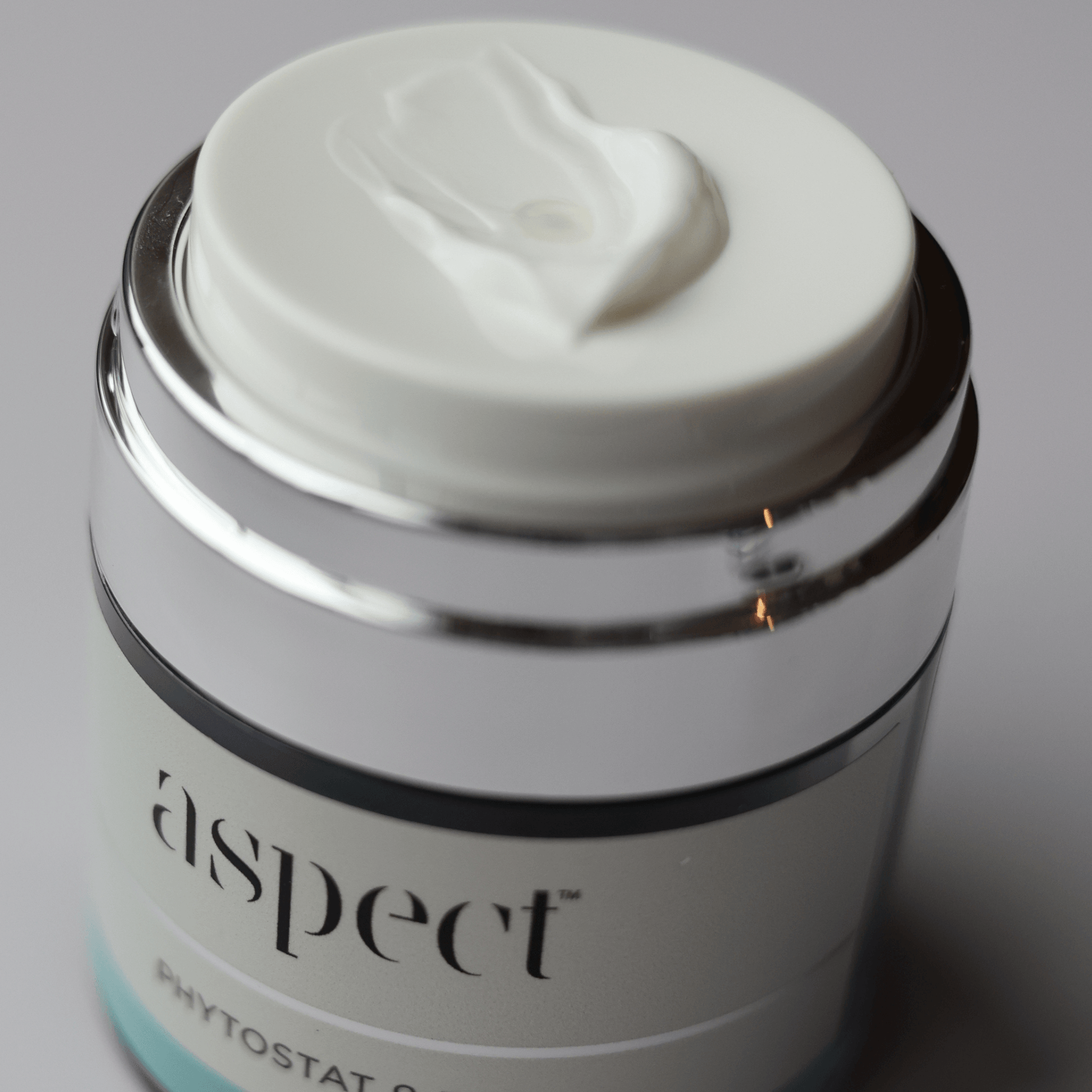 A close-up shot of Aspect Phytostat 9 cosmetic jar with a white lid showing a dollop of white cream, featuring a shiny silver metallic collar and a white label.