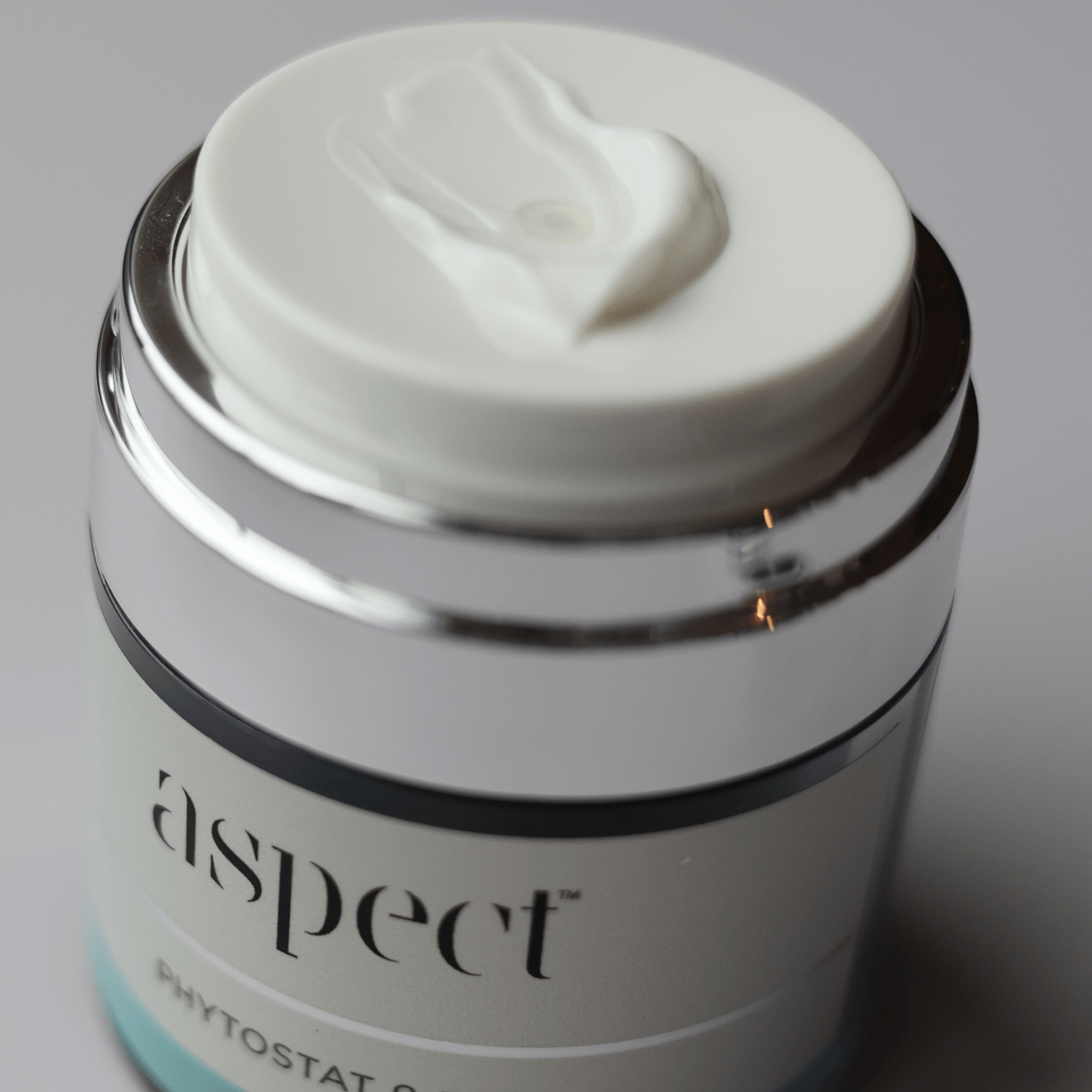 A close-up shot of Aspect Phytostat 9 cosmetic jar with a white lid showing a dollop of white cream, featuring a shiny silver metallic collar and a white label.