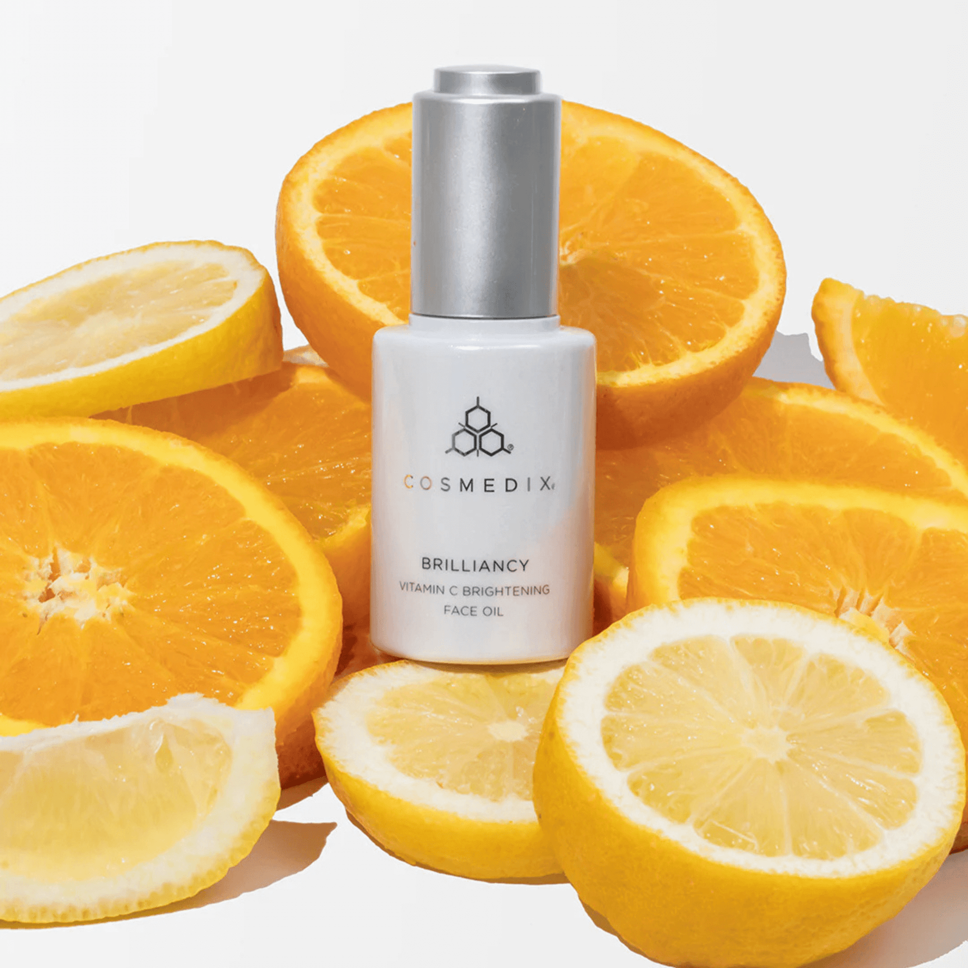 Cosmedix Brilliance skincare oil bottle surrounded by sliced oranges and lemons on a white background