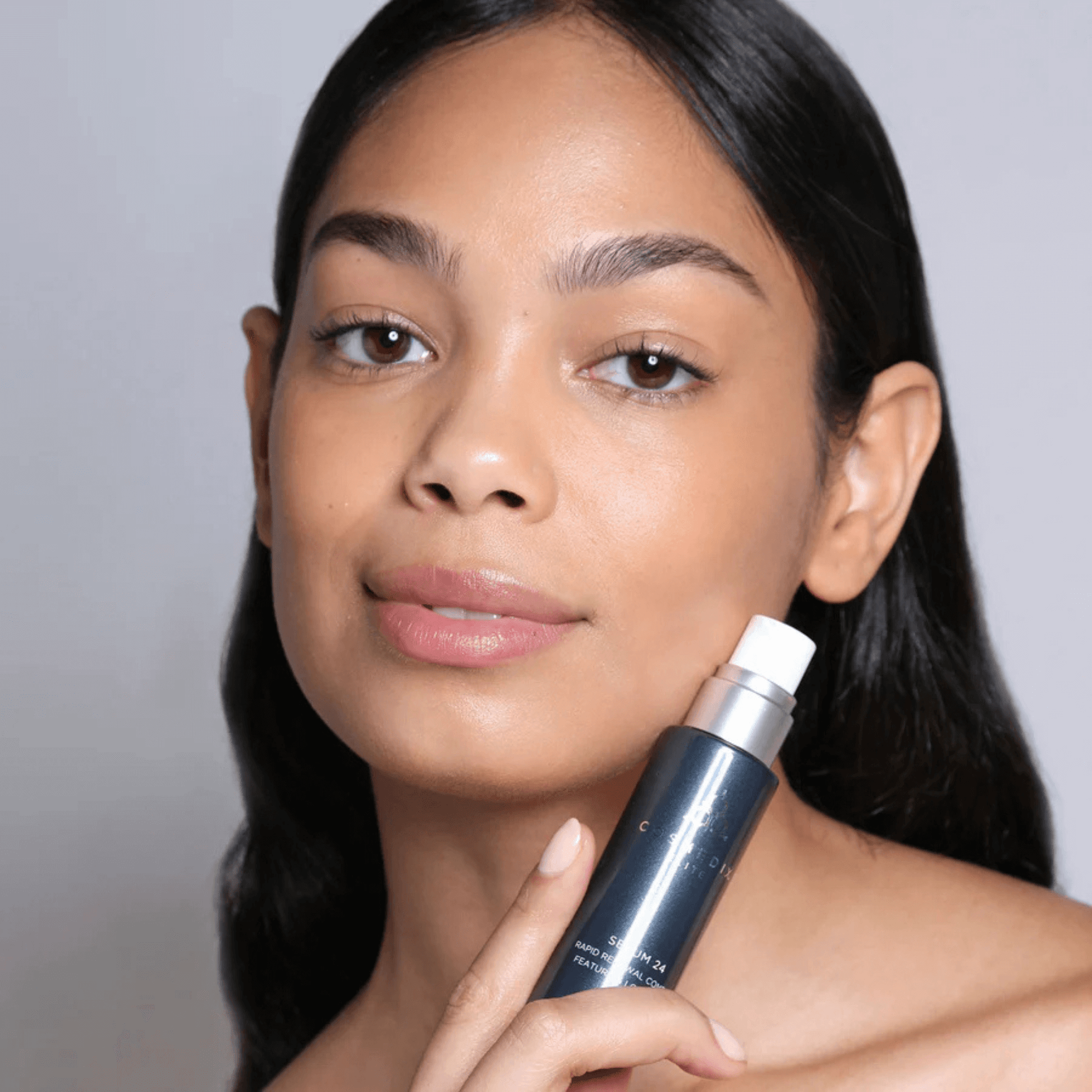 Model holding bottle of Cosmedix Elite Serum 24 against face on light grey background