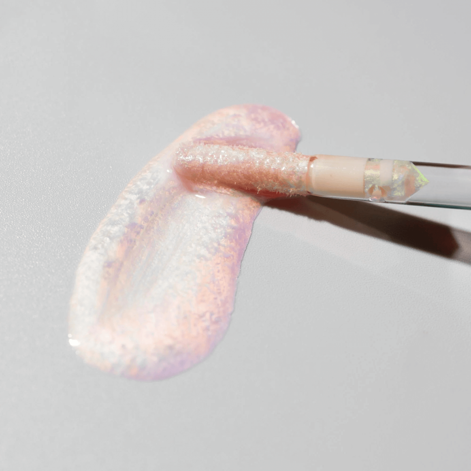 Close-up of a brush with pink and white shimmering lip gloss on a grey background