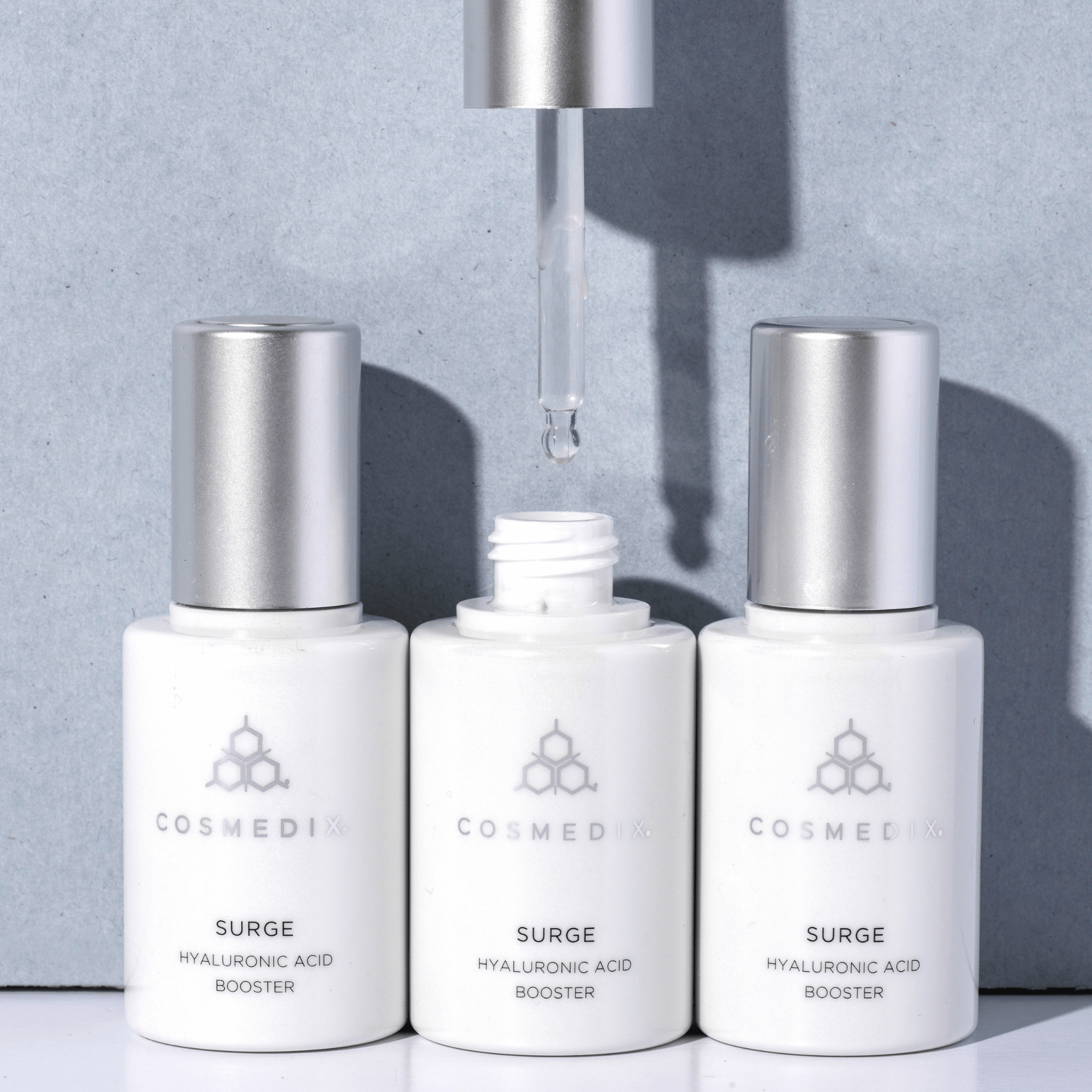 Three Cosmedix Surge Hyaluronic Acid Booster bottles on a grey background