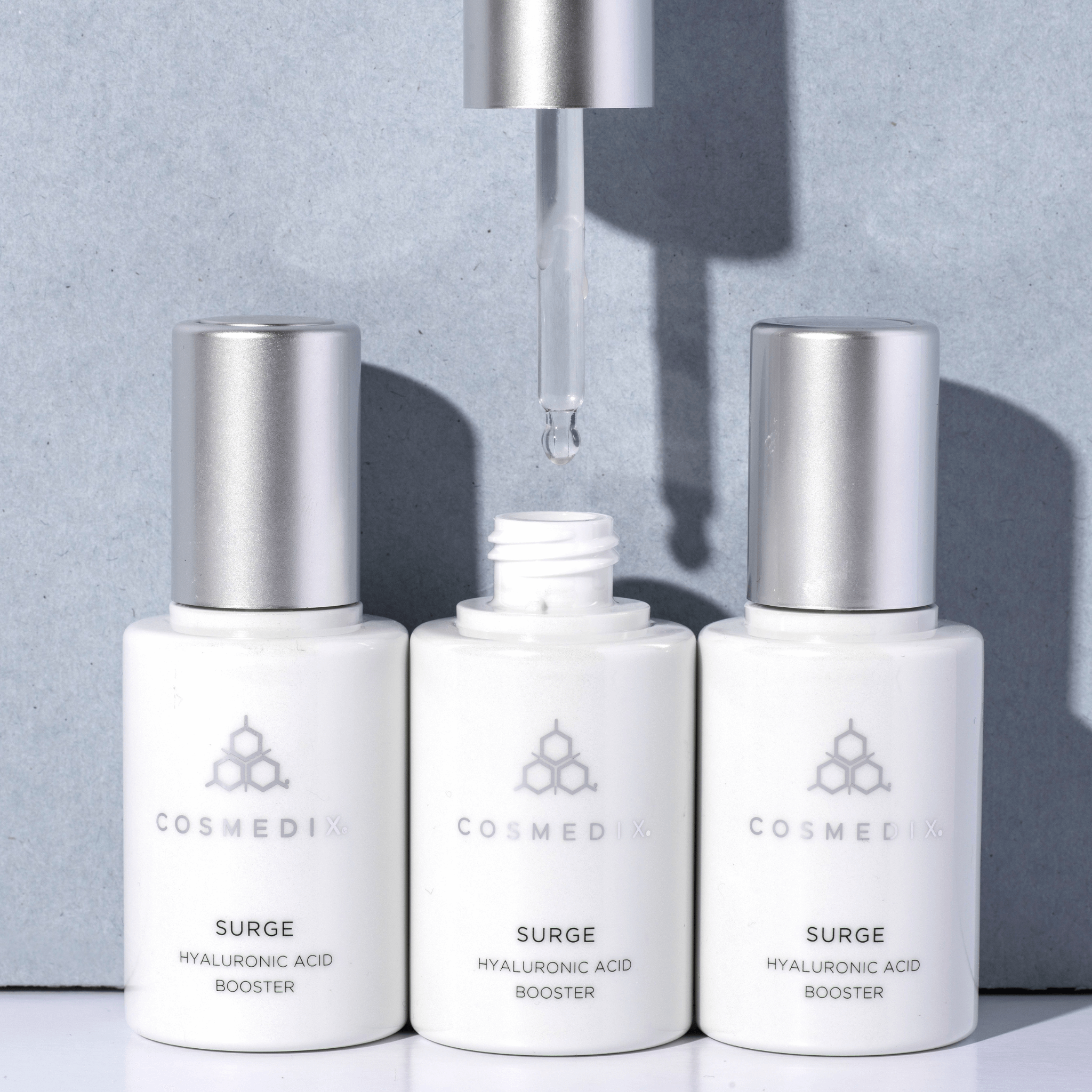 Three Cosmedix Surge Hyaluronic Acid Booster bottles on a grey background