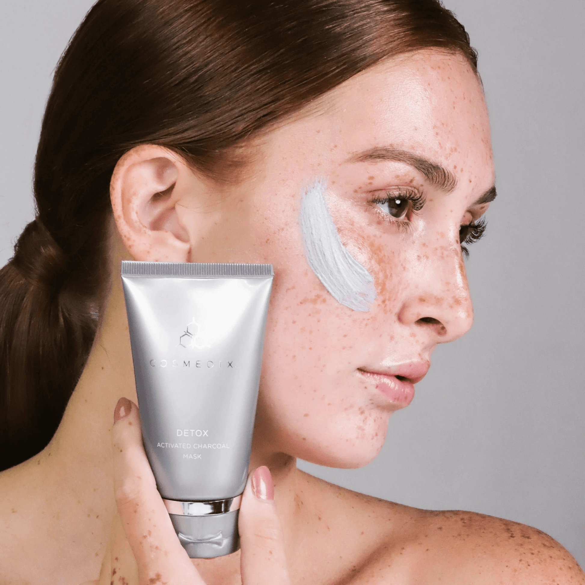 Woman applying Cosmedix Detox mask to her face with a grey background