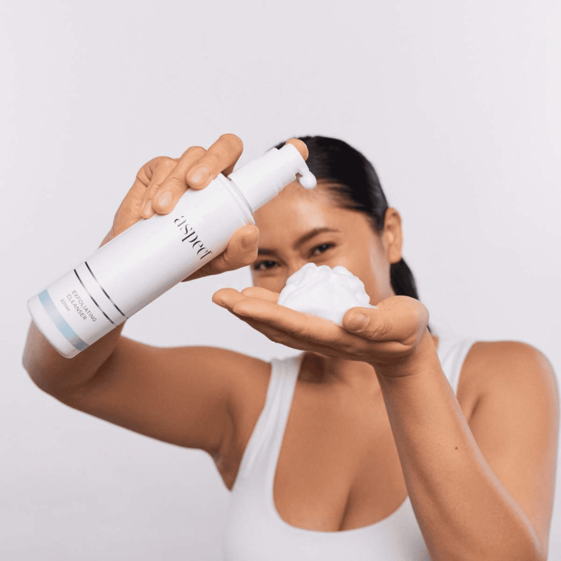 Woman holding a white bottle labeled Aspect Exfoliating Cleanser showing foam on her palm against a light grey background.