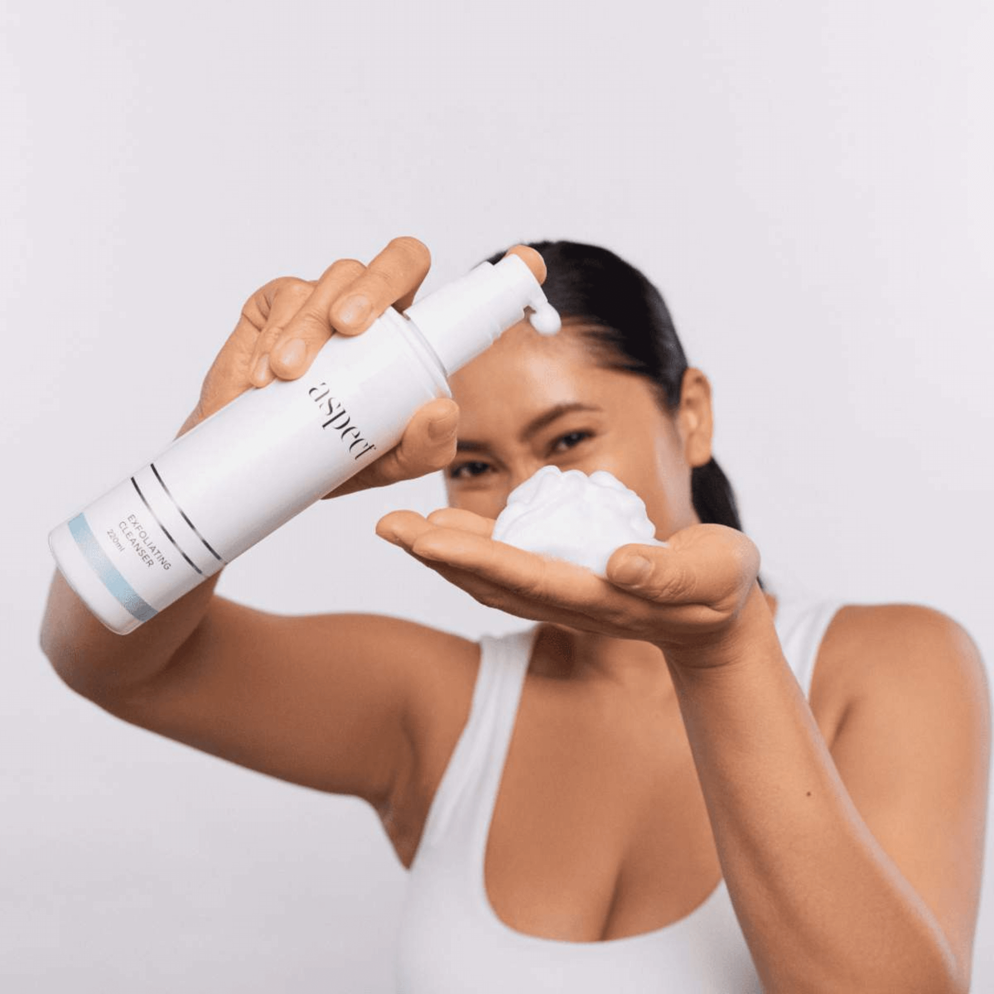 Woman holding a white bottle labeled Aspect Exfoliating Cleanser showing foam on her palm against a light grey background.