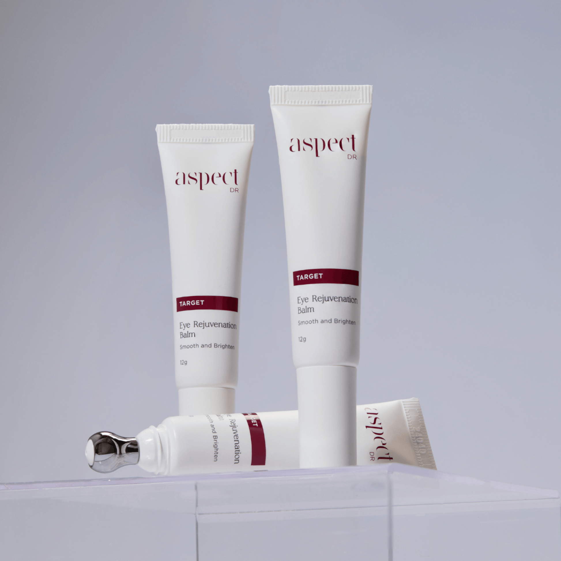 Three white cosmetic tubes of Aspect Dr Eye Rejuvenating Balm arranged on a clear acrylic block against a light grey background.