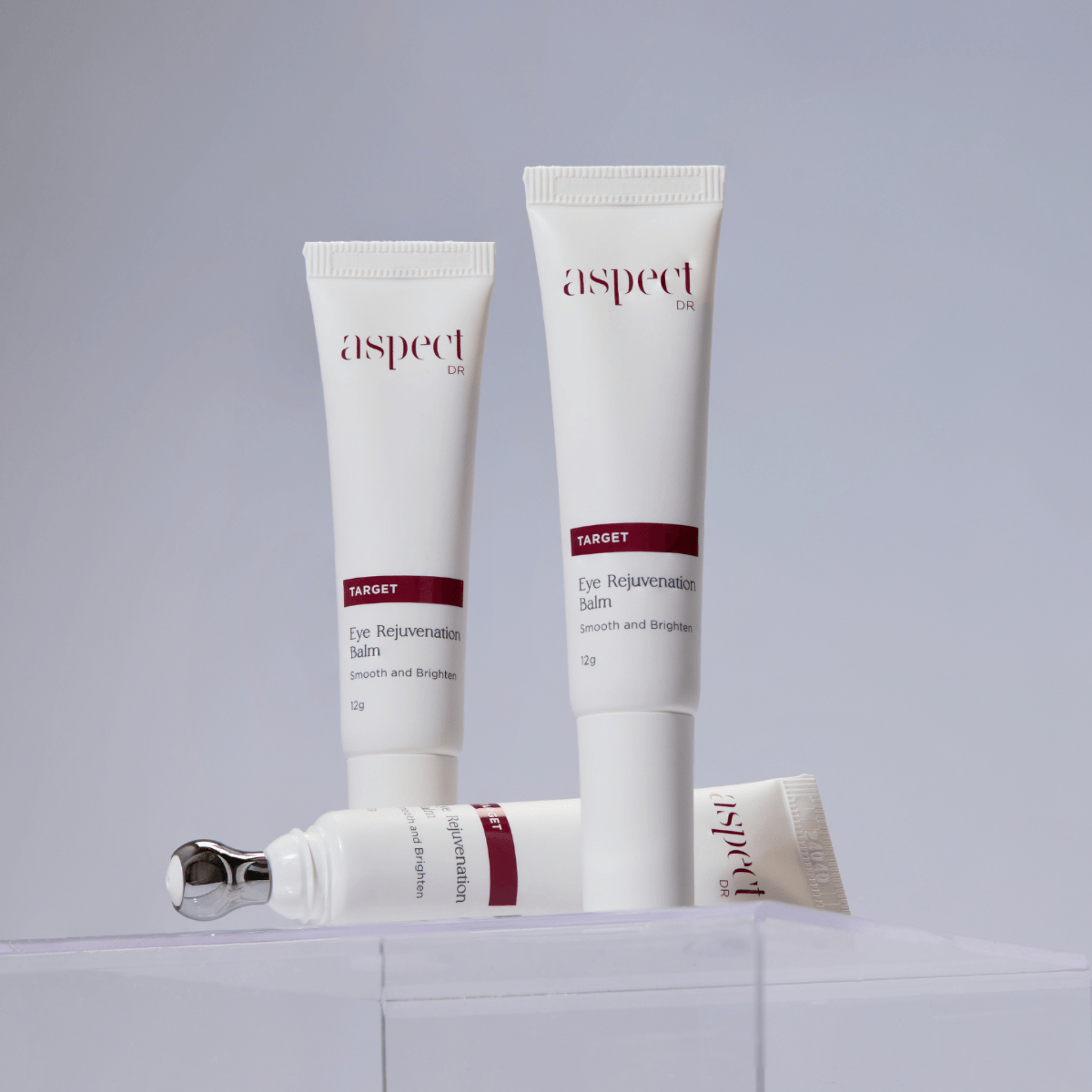 Three white cosmetic tubes of Aspect Dr Eye Rejuvenating Balm arranged on a clear acrylic block against a light grey background.