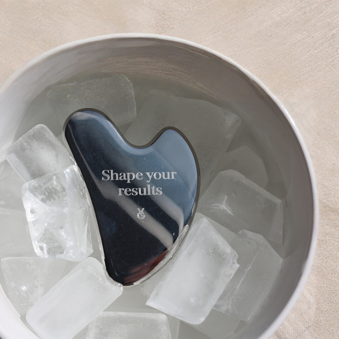 Stainless steel Gua Sha tool with 'Shape your results' text on ice cubes in a bowl.
