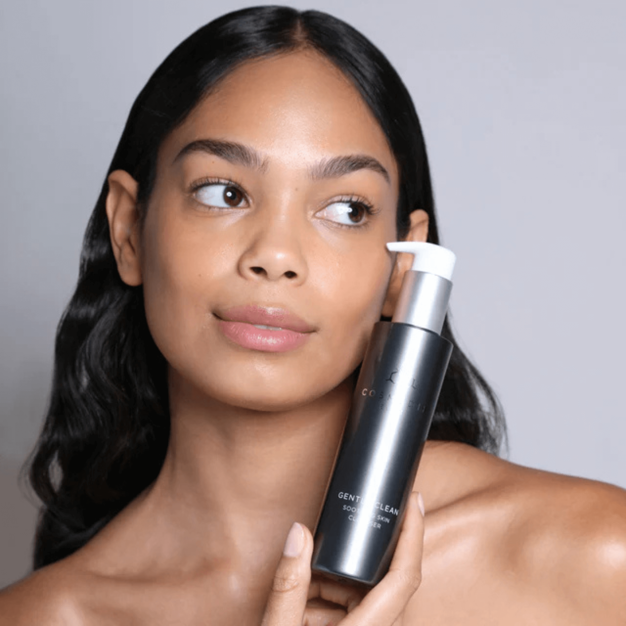 Model holding Cosmedix gentle clean cleanser bottle against face on a light grey background