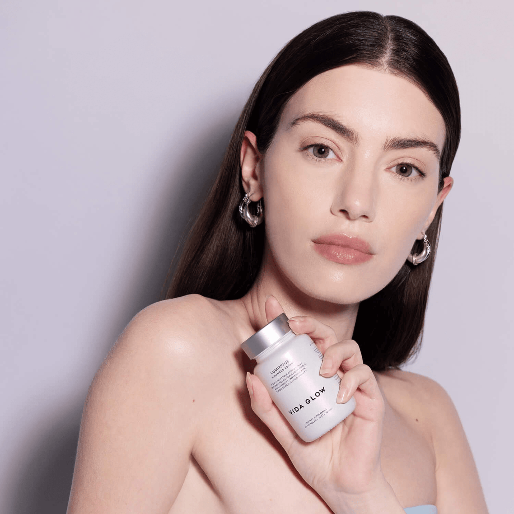 Model holding bottle of Vida Glow Luminous tablets on light purple background