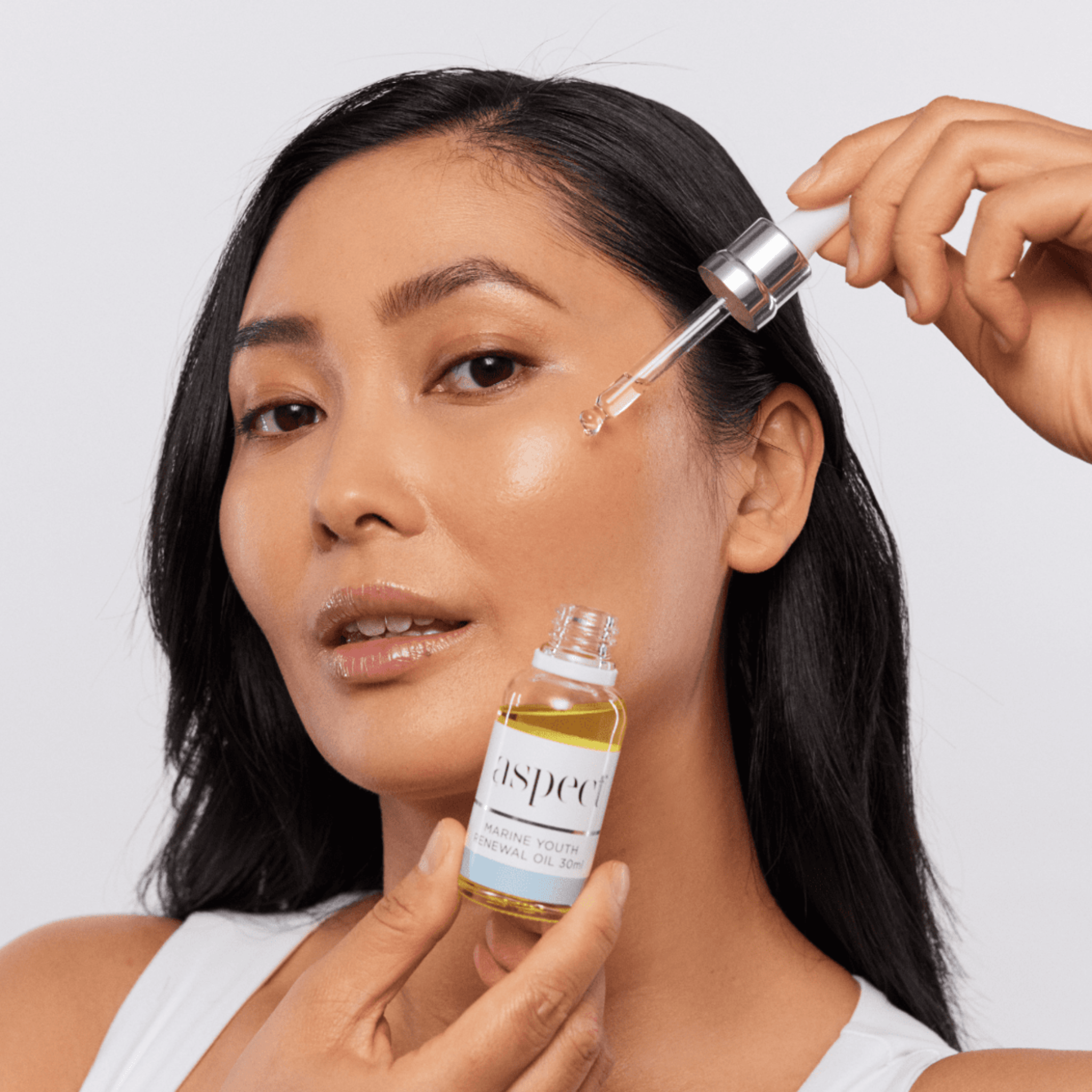 Woman applying Aspect Youth Renewal Oil skincare product to her face with a dropper and bottle