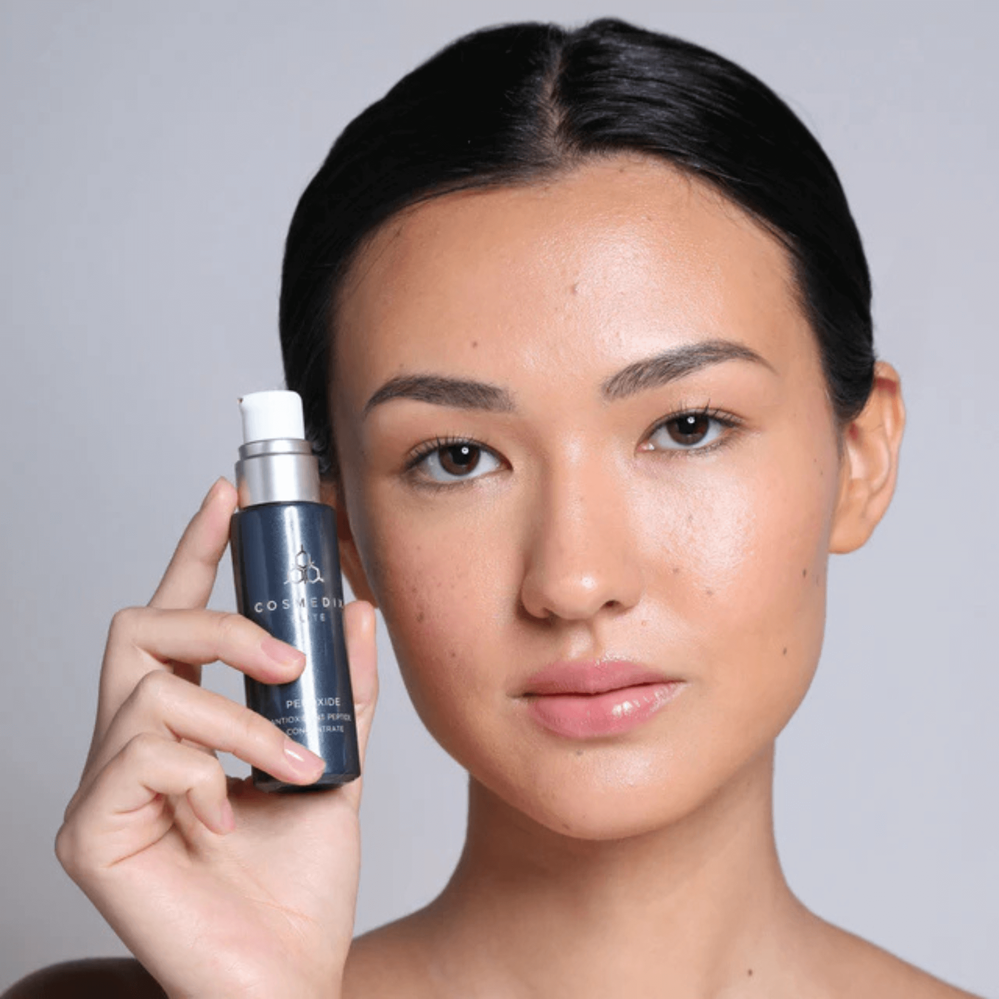 Woman holding Pepoxide COSMEDIX ELITE bottle next to face on light grey background