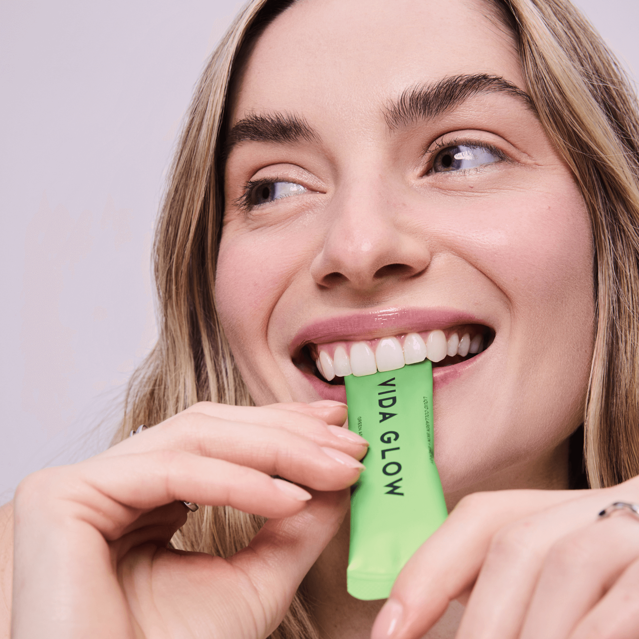 Woman squeezing a packet of Vida Glow Apple liquid collagen in her mouth against a light background