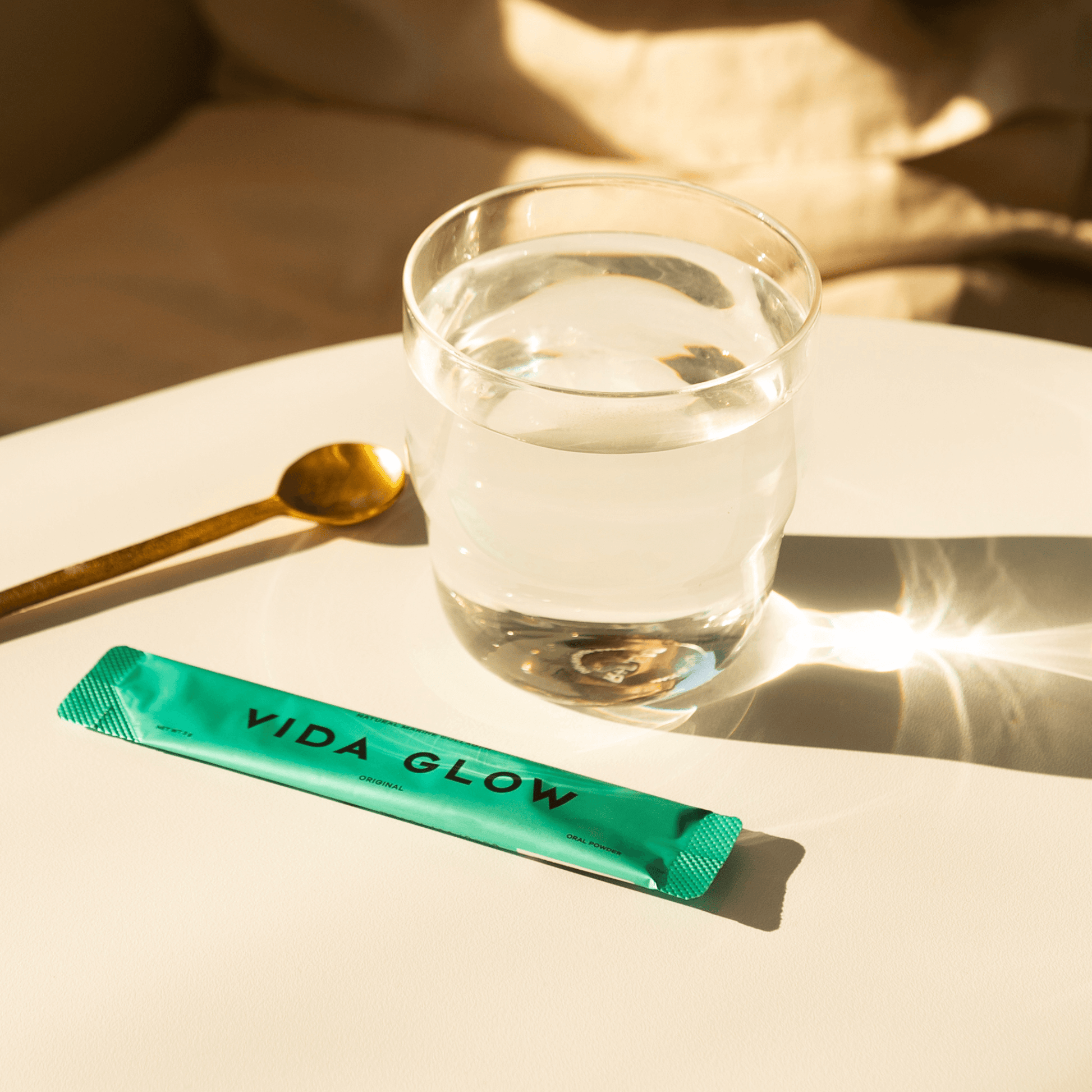 Vida Glow original flavour powder sachet sitting on table with glass of water and spoon on neutral background