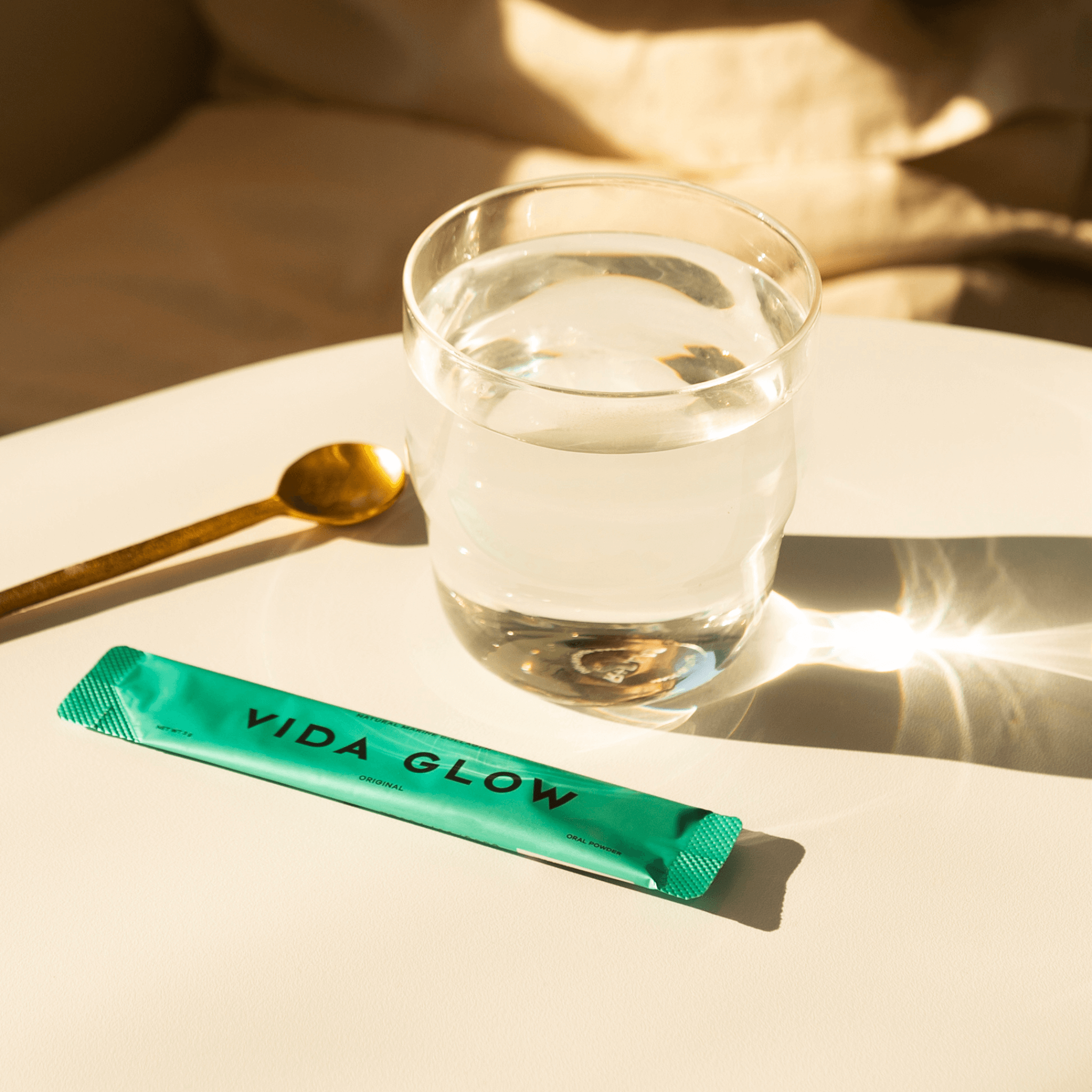 Vida Glow original flavour powder sachet sitting on table with glass of water and spoon on neutral background