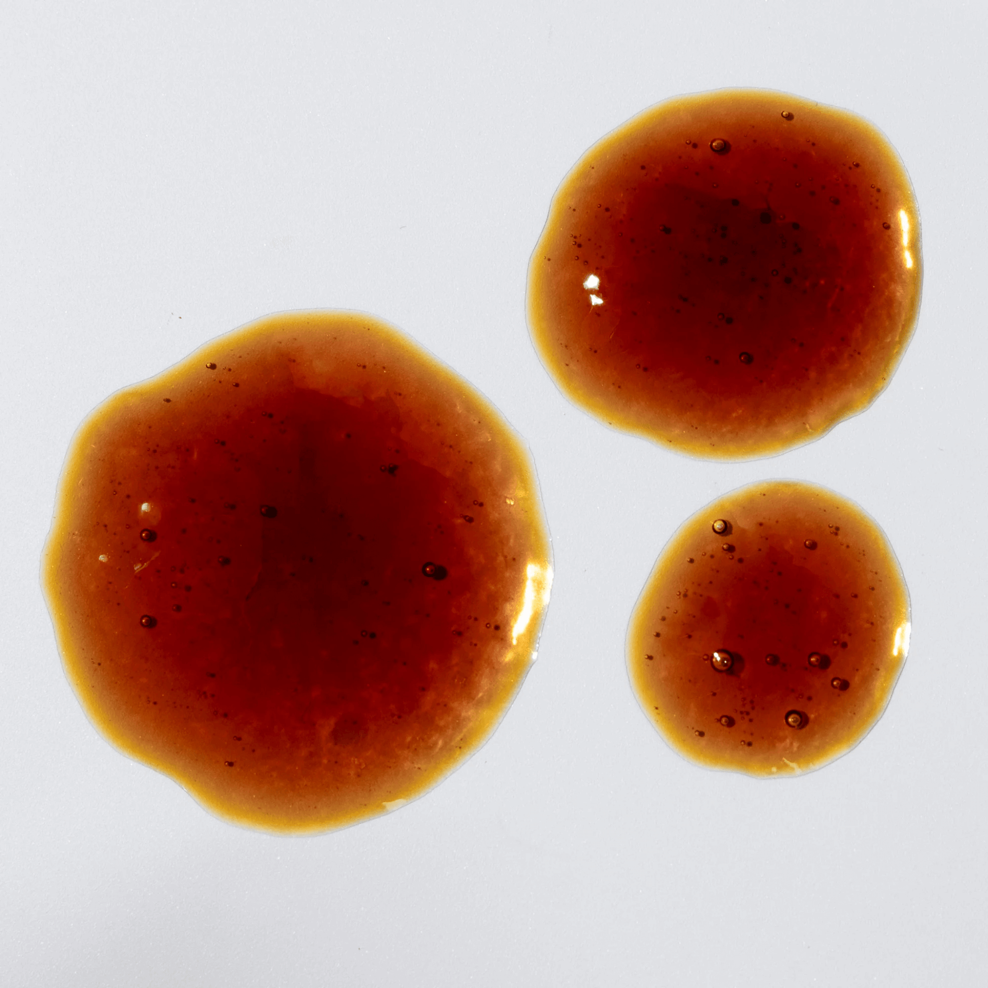 Three droplets of X-Cell+ brown liquid on a white background